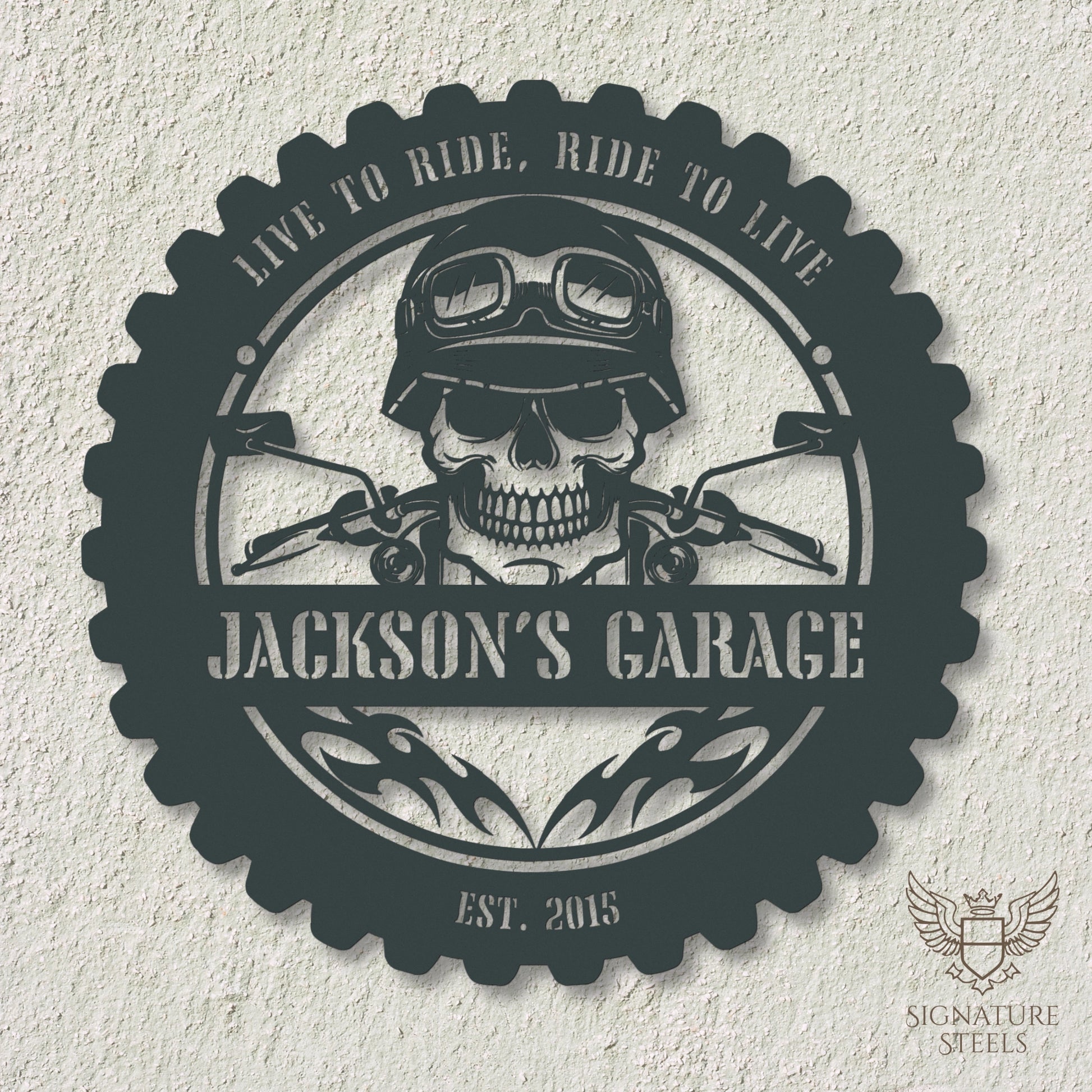 A round anthracite metal sign for a garage or man's cave with a frame imitating a motorcycle tyre, a skull wearing a biker's helmet over a motocycle handlebar and flames in the centre. The sign says 'Live to ride, ride to live", 'Jackson's Garage' and 'Est. 2015', hanging on an off-white concrete wall.