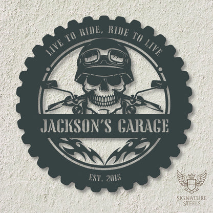 A round anthracite metal sign for a garage or man's cave with a frame imitating a motorcycle tyre, a skull wearing a biker's helmet over a motocycle handlebar and flames in the centre. The sign says 'Live to ride, ride to live", 'Jackson's Garage' and 'Est. 2015', hanging on an off-white concrete wall.
