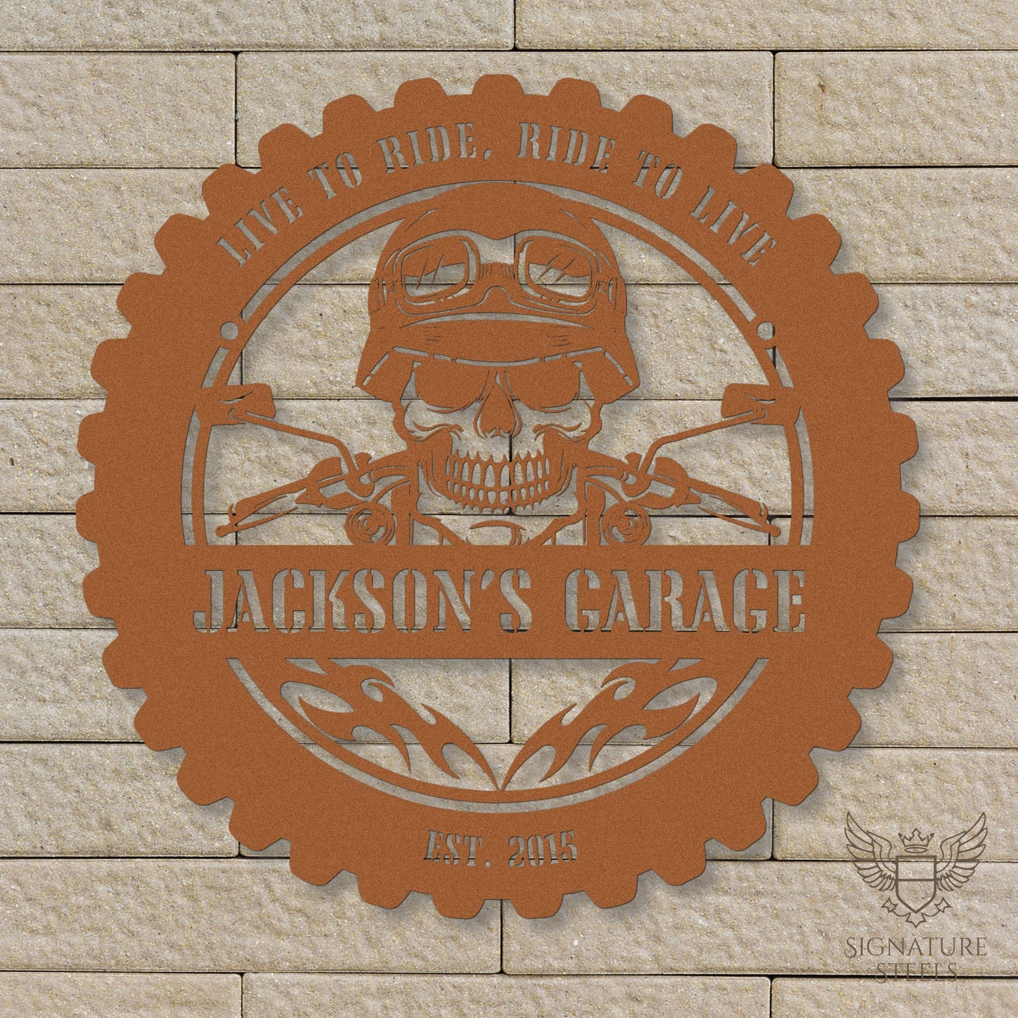 A round copper metal sign for a garage or man's cave with a frame imitating a motorcycle tyre, a skull wearing a biker's helmet over a motocycle handlebar and flames in the centre. The sign says 'Live to ride, ride to live", 'Jackson's Garage' and 'Est. 2015', hanging on a beige brick wall.