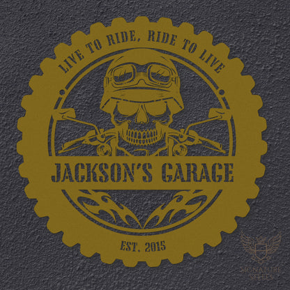 A round gold metal sign for a garage or man's cave with a frame imitating a motorcycle tyre, a skull wearing a biker's helmet over a motocycle handlebar and flames in the centre. The sign says 'Live to ride, ride to live", 'Jackson's Garage' and 'Est. 2015', hanging on a dark grey concrete wall.