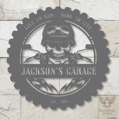 A round silver metal sign for a garage or man's cave with a frame imitating a motorcycle tyre, a skull wearing a biker's helmet over a motocycle handlebar and flames in the centre. The sign says 'Live to ride, ride to live", 'Jackson's Garage' and 'Est. 2015', hanging on a marble-imitating brick wall.