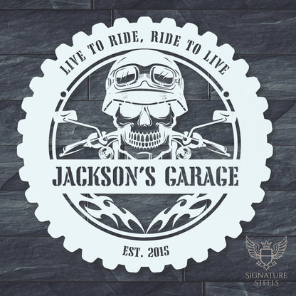 A round white metal sign for a garage or man's cave with a frame imitating a motorcycle tyre, a skull wearing a biker's helmet over a motocycle handlebar and flames in the centre. The sign says 'Live to ride, ride to live", 'Jackson's Garage' and 'Est. 2015', hanging on a dark grey brick wall.