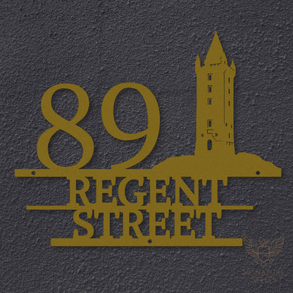 Gold metal address sign with house number 89, street name Regent Street, horizontal parallel lines and a silhouette of Scrabo Tower, a landmark of Newtownards in Northern Ireland, hanging on a dark grey exterior wall.