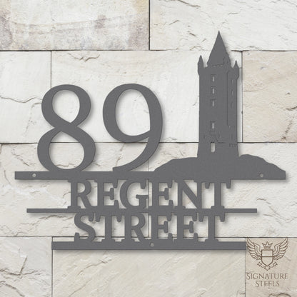 Silver metal address sign with house number 89, street name Regent Street, horizontal parallel lines and a silhouette of Scrabo Tower, a landmark of Newtownards in Northern Ireland, hanging on a white and beige brick wall.